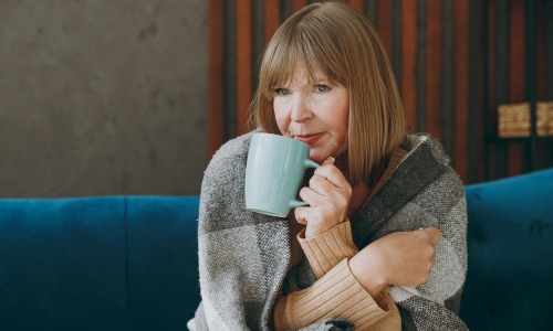Elderly sad woman 50s years old wrap in plaid sits on sofa drink tea stay at home flat spending free spare time in living room indoor Healthy lifestyle ill sick disease treatment cold season concept